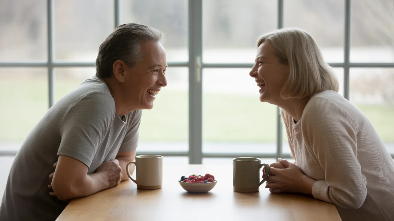 Two seniors are laughing together while sitting at a kitchen table with coffee and a bowl of berries in soft, natural light.