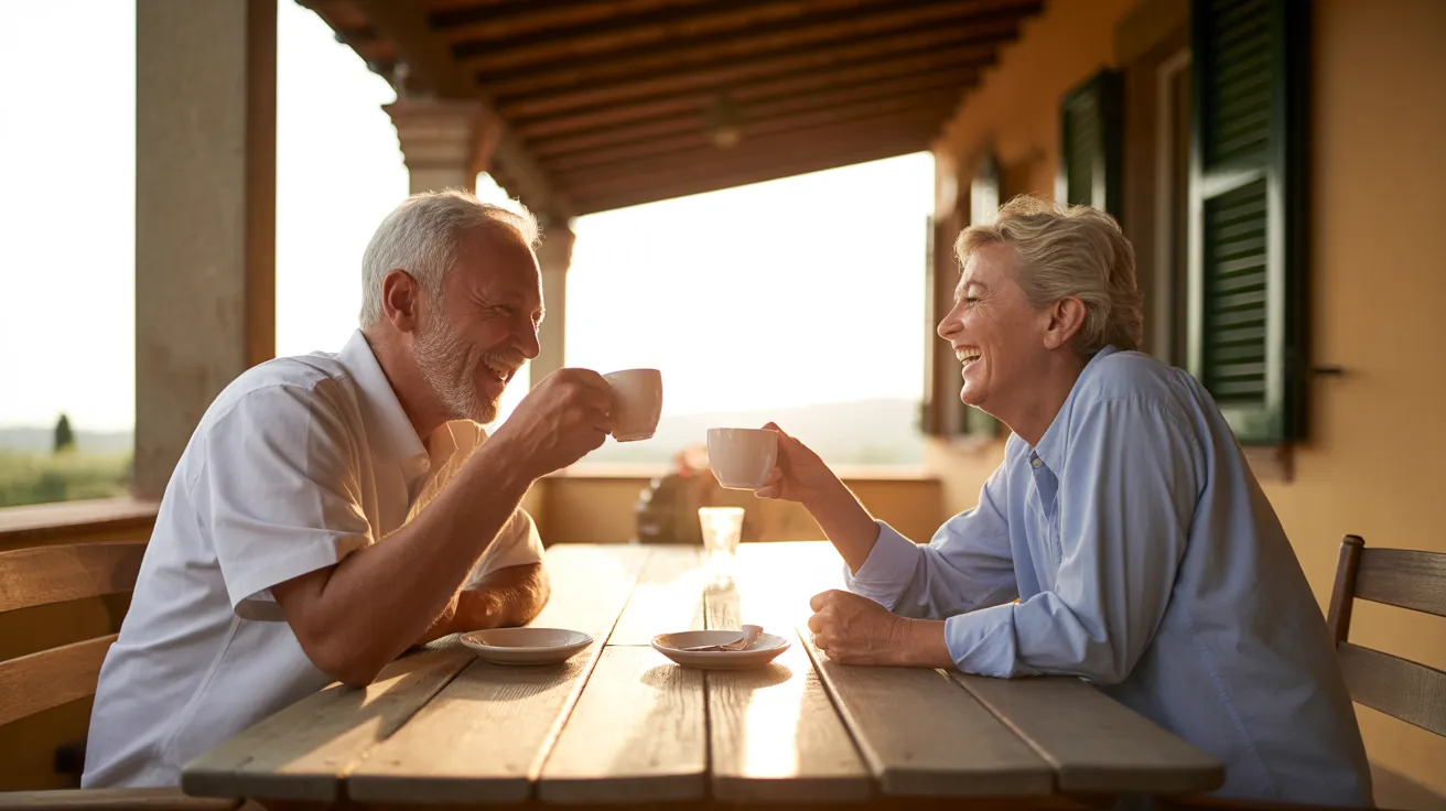 A senior couple laughs over coffee at a sunlit table on a patio under a soft, overcast sky.