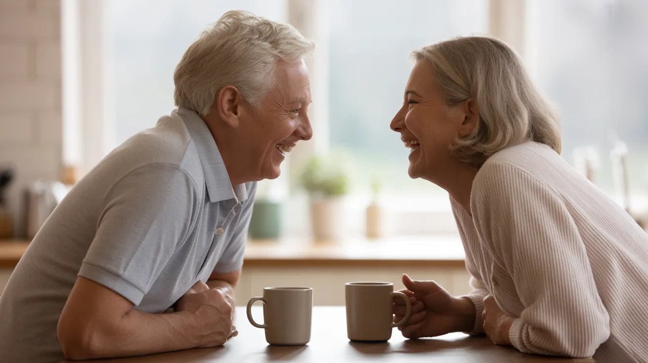 A senior man and woman are laughing together while sitting opposite each other at a kitchen table with coffee mugs.