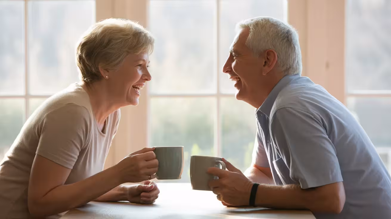 A senior man and woman sit opposite each other at a kitchen table, laughing together over coffee, in a brightly lit room.