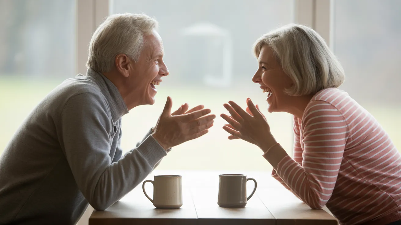 A senior man and woman laugh together while sitting at a bright kitchen table with coffee mugs.
