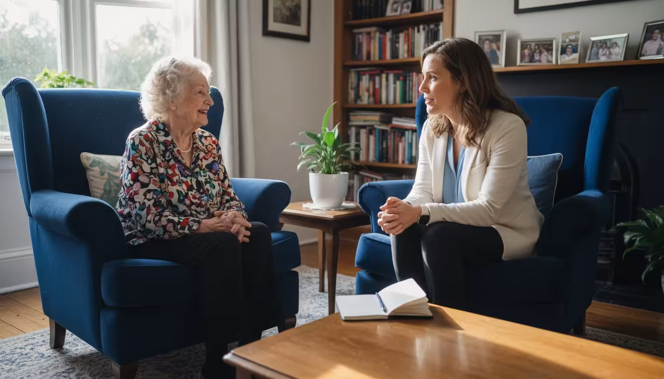An older woman and a female caregiver sit in armchairs in a living room, looking at each other with warm smiles as they converse.