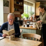 A retired husband sitting at a kitchen table while his working wife packs her bag for work in the background of their sunlit home.